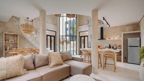 Un intérieur moderne avec un canapé beige, une table basse ronde et un coin cuisine équipé d’un réfrigérateur et de tabourets de bar.