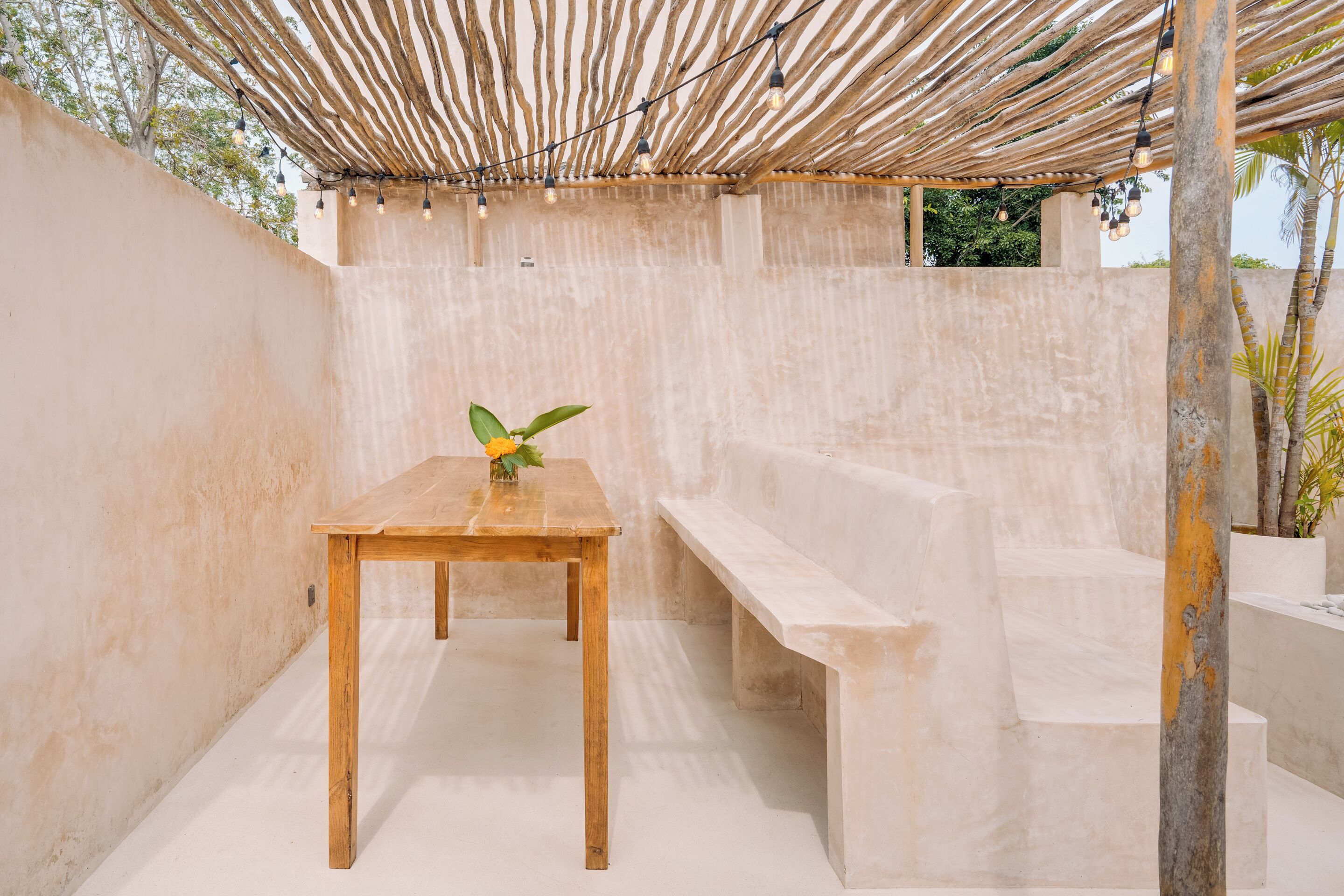 Un patio al aire libre con una mesa y banco de madera, luces colgantes y un muro de hormigón.