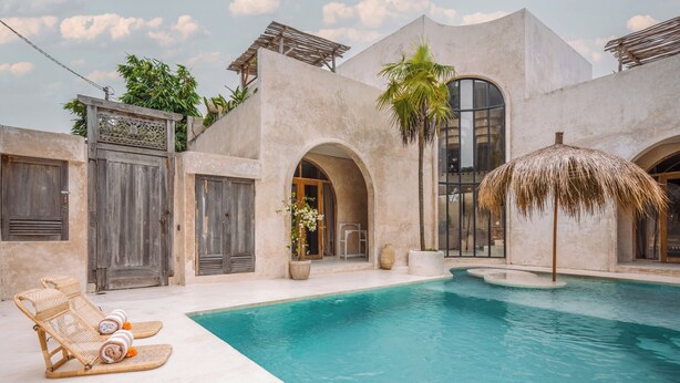 Un espace piscine aménagé avec des chaises longues, un parasol en chaume et un bâtiment doté d’arcades et de grandes fenêtres.