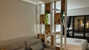 Un salon moderne avec un canapé gris, des chaises en bois, une table blanche et un lit recouvert d’une couette blanche. On y trouve également une bibliothèque en bois agrémentée de plantes et d’objets décoratifs.