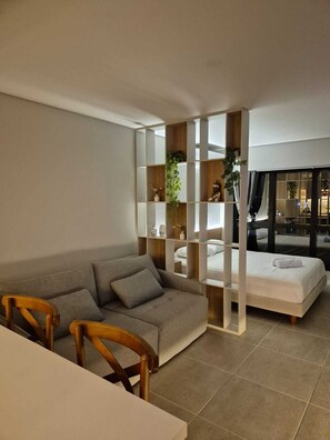 A modern living room with a grey sofa, wooden chairs, a white table, and a bed with a white duvet. There is a wooden bookshelf with plants and decorative items. - High-end and cozy studio apartment with a pool and close to the beaches. (Florianópolis)