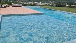 Une piscine d’un bleu éclatant, dotée d’une terrasse en bois et d’un espace détente, le tout entouré de collines verdoyantes et sous un ciel dégagé.