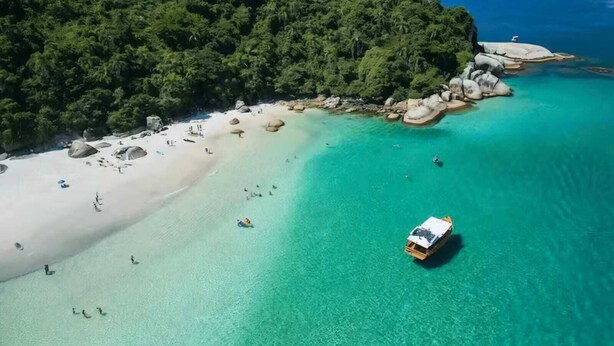 Vue aérienne d’une plage aux eaux turquoise et limpides, au sable blanc et aux affleurements rocheux.