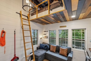 A loft-style room with a loft bed, a ladder, a couch with cushions, and a broom.