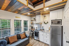 A modern kitchen with white cabinets, a stainless steel refrigerator, and a built-in microwave. There is a gray sofa with brown cushions and a window with a view of greenery.