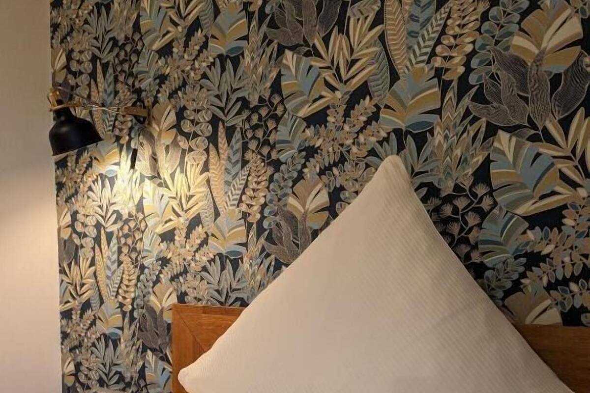 A neatly made bed with white linens and a single pillow, set against a wall with a detailed floral wallpaper pattern.