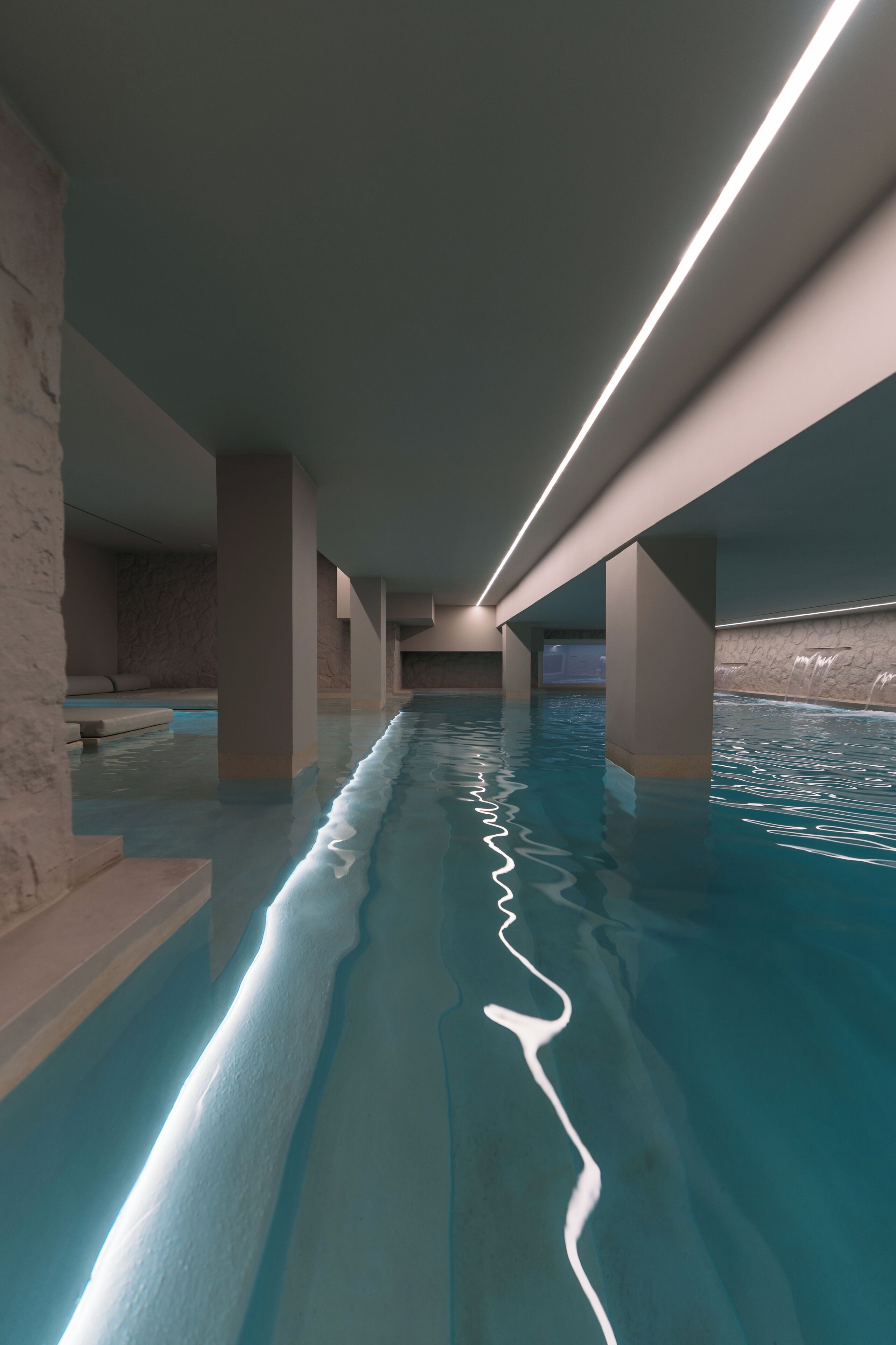 An indoor swimming pool with clear water, supported by columns, and illuminated by overhead lighting.
