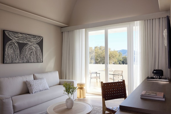A modern living room with a sofa, a coffee table with a plant, a dining area with a chair, and a view through large windows. - Santorini Heart Hotel & Spa (Santorini)