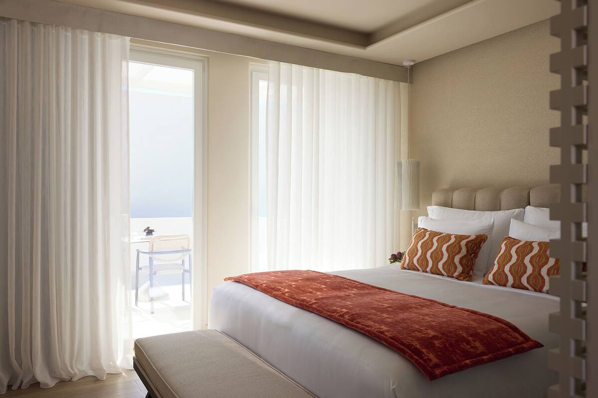 A hotel room with a bed, a bench, and a view of the outdoors.