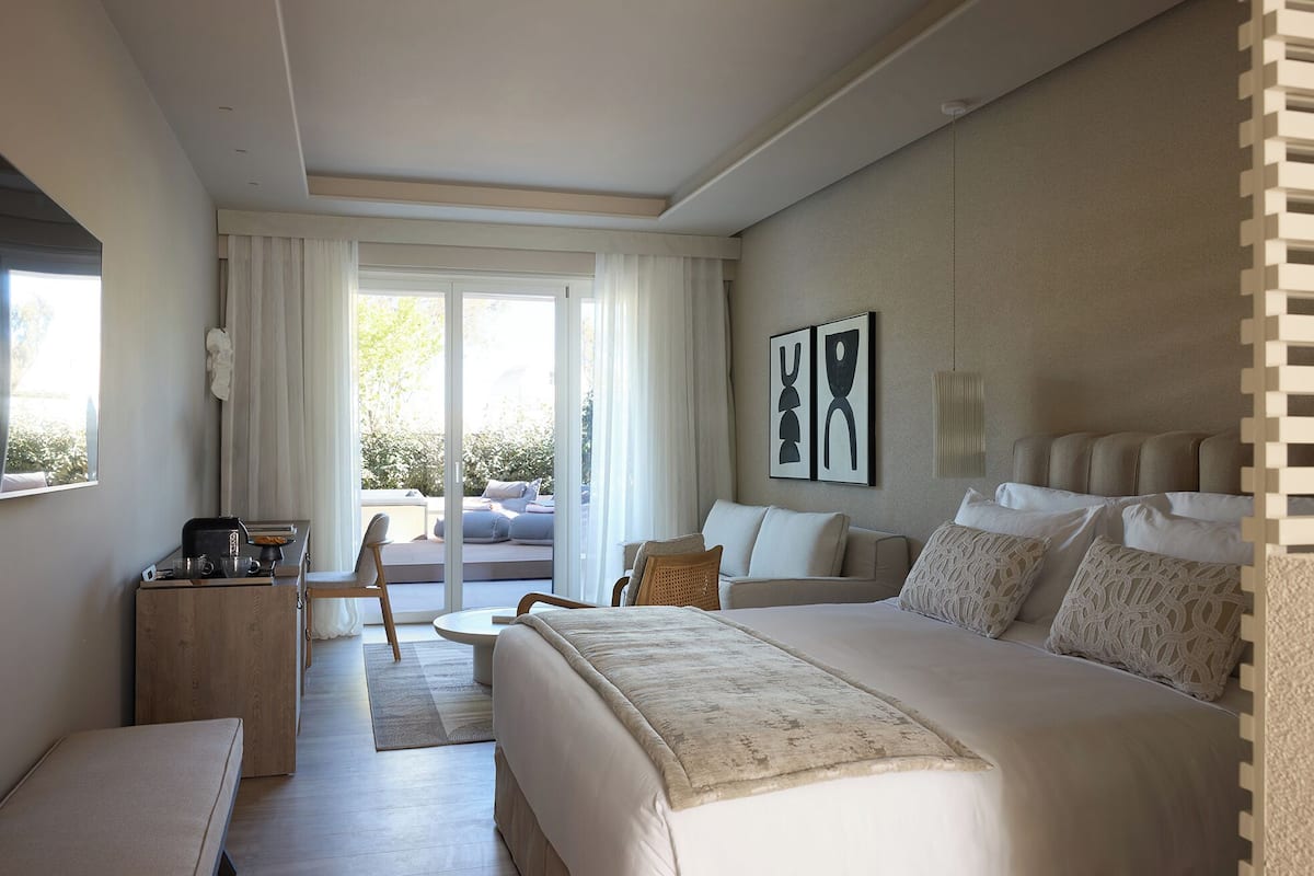 A hotel room with a bed, a seating area with a table and chairs, and a view through a large window.
