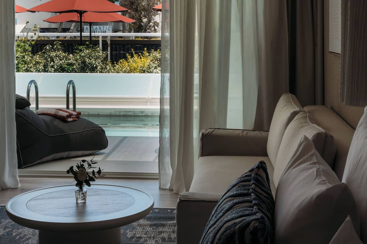 A modern hotel room with a sofa, a round coffee table, and a view of an outdoor pool area.