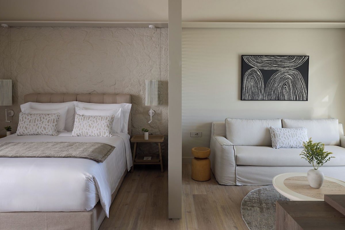 A bedroom with a bed, bedside tables, and a nightstand. There is a living area with a sofa, a coffee table, and a wall-mounted artwork.