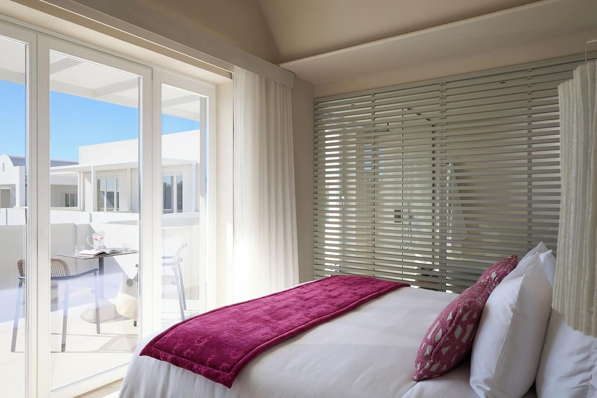 A bedroom with a bed, white curtains, and a view of a balcony with a table and chairs.
