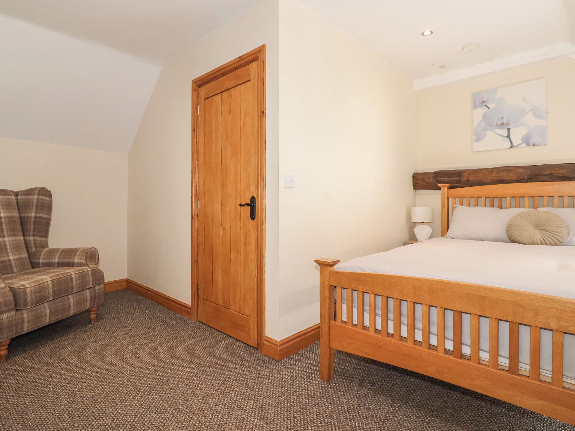 A bedroom with a wooden bed, a plaid chair, a wooden door, and a painting on the wall.