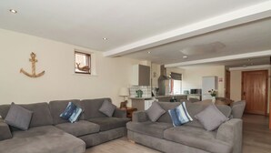 Ein geräumiger Wohnbereich mit zwei grauen Sofas, Holzboden, einer Küche im Hintergrund und einer Holztür.