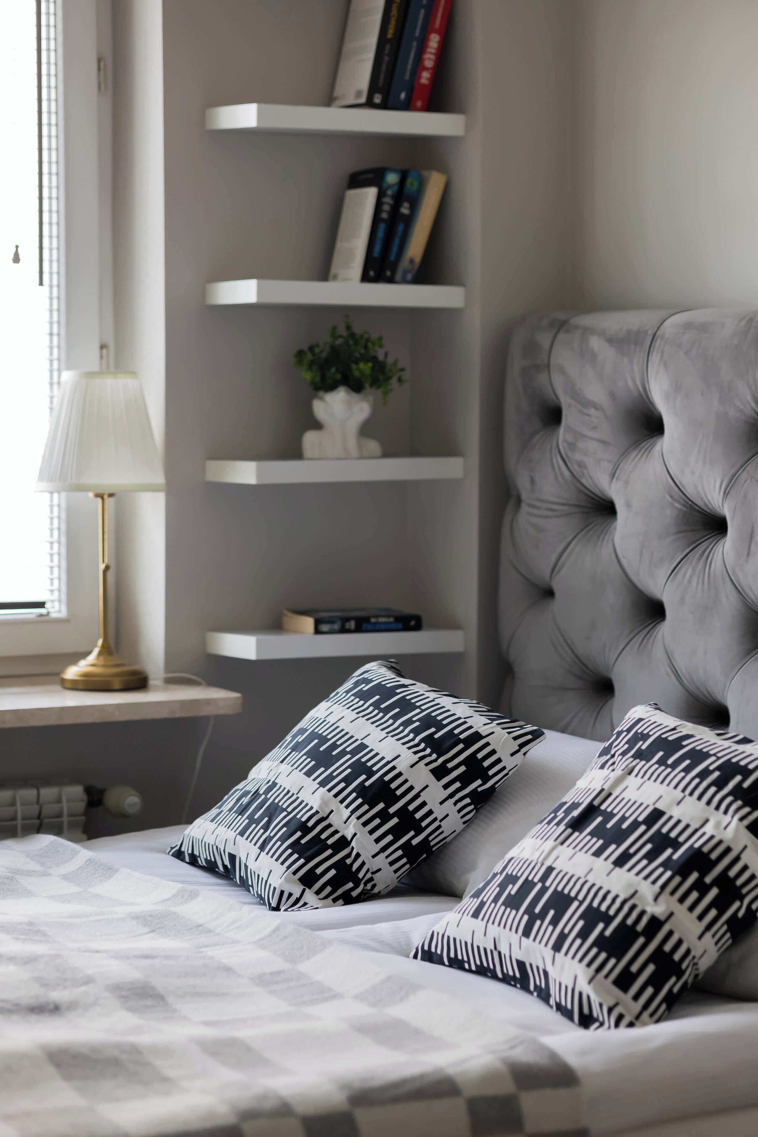A bedroom with a tufted headboard, a bedside table with a lamp, and a bookshelf with books and decorative items.