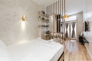 Ein Schlafzimmer mit Bett, einer Wandlampe, Holzboden und Blick ins Freie durch ein Fenster.