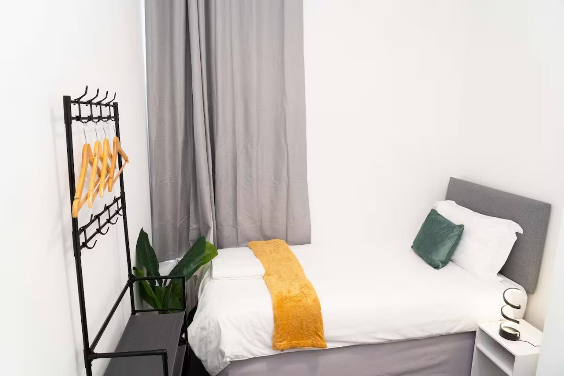 A neatly arranged hotel room with a bed, a side table, a plant, and a black metal rack with wooden items.