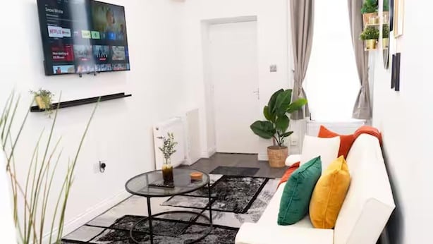 A living room with a flat-screen TV mounted on the wall, a white sofa with colorful cushions, a glass coffee table, and a potted plant. - Near Transport - Parking - Wifi - Sleeps 3 (Newcastle-upon-Tyne)