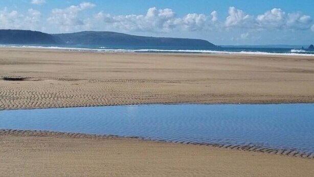 Una playa con una extensa franja de arena, un mar tranquilo y montañas lejanas bajo un cielo despejado.