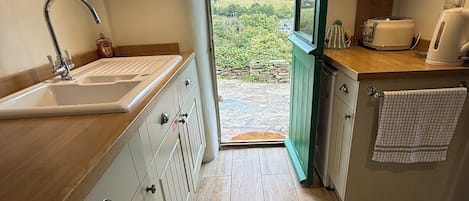 A kitchen with a sink, wooden cabinets, a green door leading outside, and a view of the countryside.