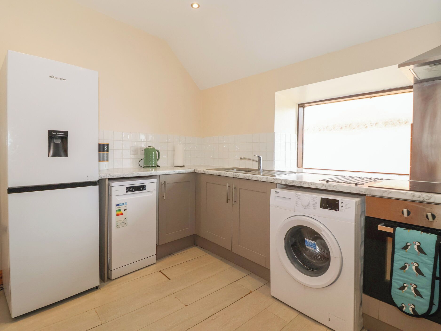 A compact kitchen with a refrigerator, dishwasher, washing machine, and a window providing natural light.