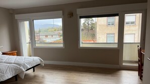 Un dormitorio con cama, suelo de madera y ventanas amplias con vistas a edificios y árboles.