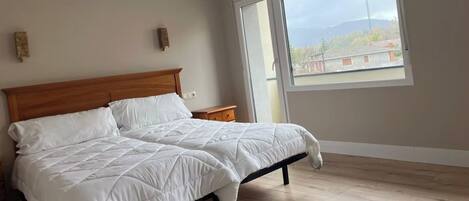 A bedroom with a wooden bed, white bedding, a nightstand, and a large window with a view of buildings and a tower.