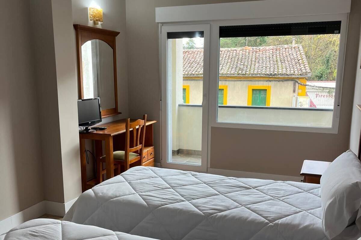 Ein Schlafzimmer mit zwei Betten, einem Schreibtisch, einem Stuhl, einem Spiegel und einem Fenster mit Blick auf ein Gebäude.