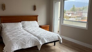Un dormitorio con cama de madera, mesita de noche y ventana grande con vista a edificios y árboles.