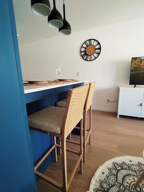 A dining area with a blue wall, a white table, and two wicker chairs. A black clock is on the wall, and a TV is on a white cabinet. - Mía Bonita - urban oasis with private terrace (Montevideo)