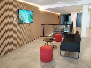 Un interior moderno con un televisor montado en una pared de madera, barandilla de vidrio, escalera y una zona de estar con sofá y reposapiés.
