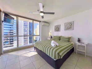 Ein Hotelzimmer mit Bett, Deckenventilator, Nachttisch, Statue und einem großen Fenster mit Blick auf die Stadt.