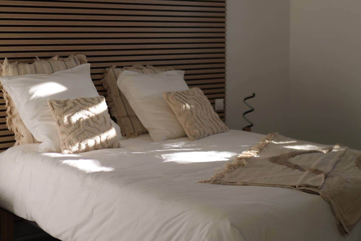 Een bed met witte beddengoed en meerdere kussens, een houten lattenbalken hoofdbord en een effen muur.