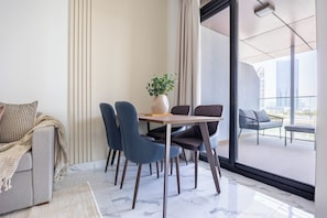 A modern living room with a sofa, a dining table with chairs, and a balcony with a view of the city. - Bloomfields Vista Elegance 1Br Al Maryah (Abu Dhabi)
