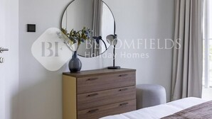 Ein Schlafzimmer mit einem Bett, einer Kommode mit Schubladen, einem runden Spiegel und einer Vase mit Blumen.