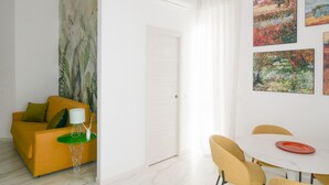 A modern living room with a sofa, a small table with a lamp, and a dining area with chairs and a table.