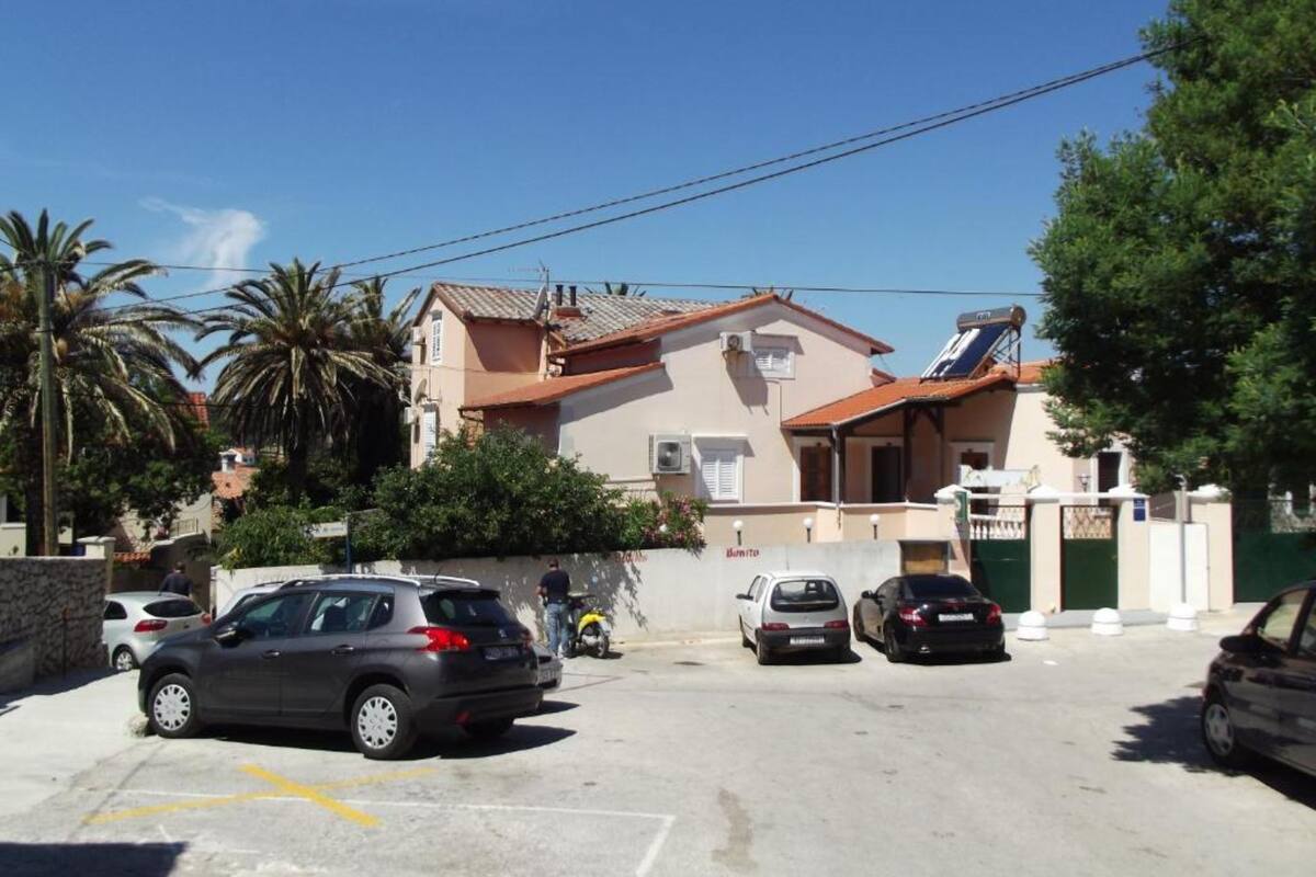 A parking area with several cars, a building with a tiled roof, and a palm tree in the background.