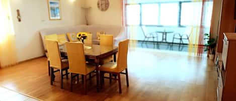 A dining area with a wooden table and chairs, a clock on the wall, and a view of the outside through large windows.