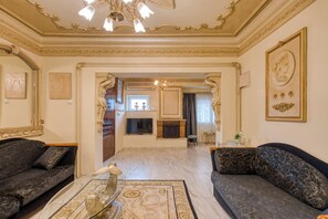 A spacious living room with two dark sofas, a glass coffee table, and a fireplace. The room features ornate gold detailing on the ceiling and walls, and a chandelier.