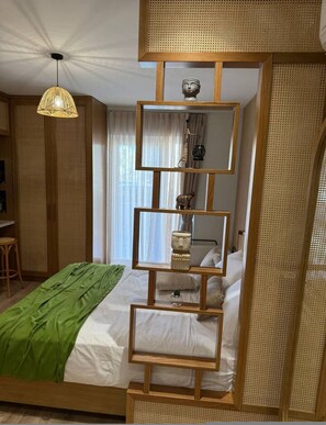 A bedroom with a bed, a green blanket, a hanging light fixture, and a wooden shelf with decorative items. - Luvra 1 Dh rmi Spacious Balcony Hideaway (Himarë)