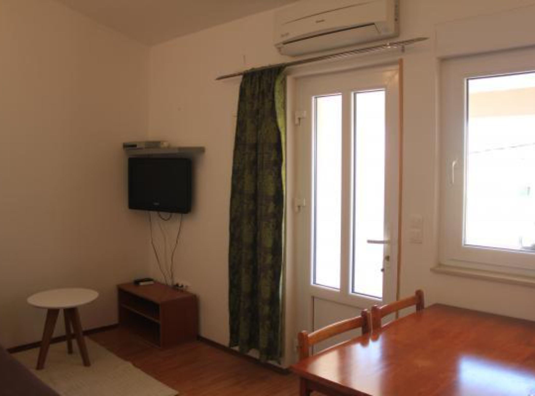 Una habitación con una mesa de comedor de madera, un televisor montado en la pared, un mueble de madera pequeño y un ventanal con cortina corredera.