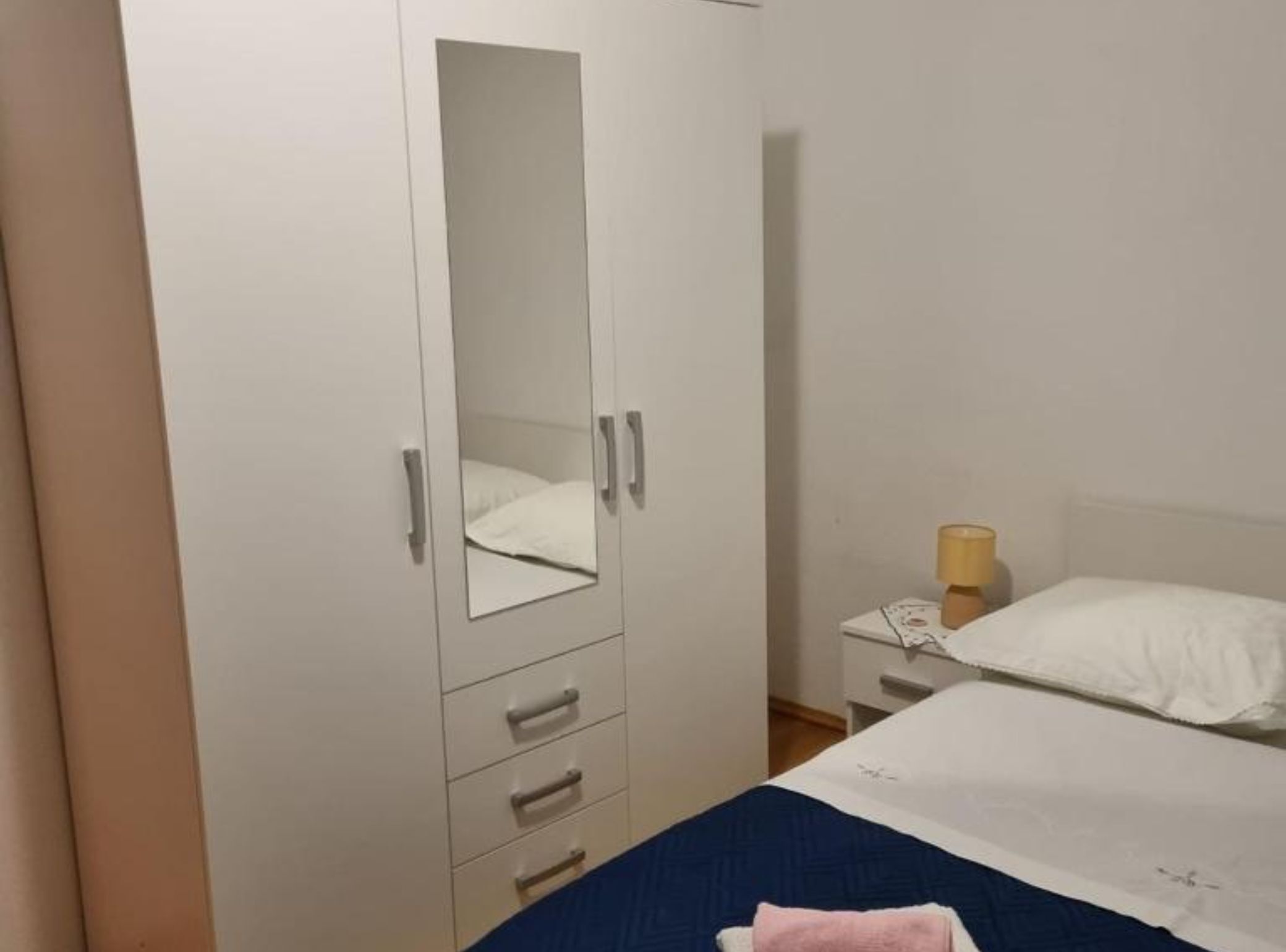 Un dormitorio con cama, mesita de noche con lámpara, armario con espejo y cómoda con cajones.