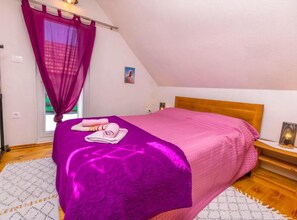 A bedroom with a bed, a window with purple curtains, a bedside table with a lamp, and a wall-mounted picture. - Holiday Home Kuzmino (Primosten)