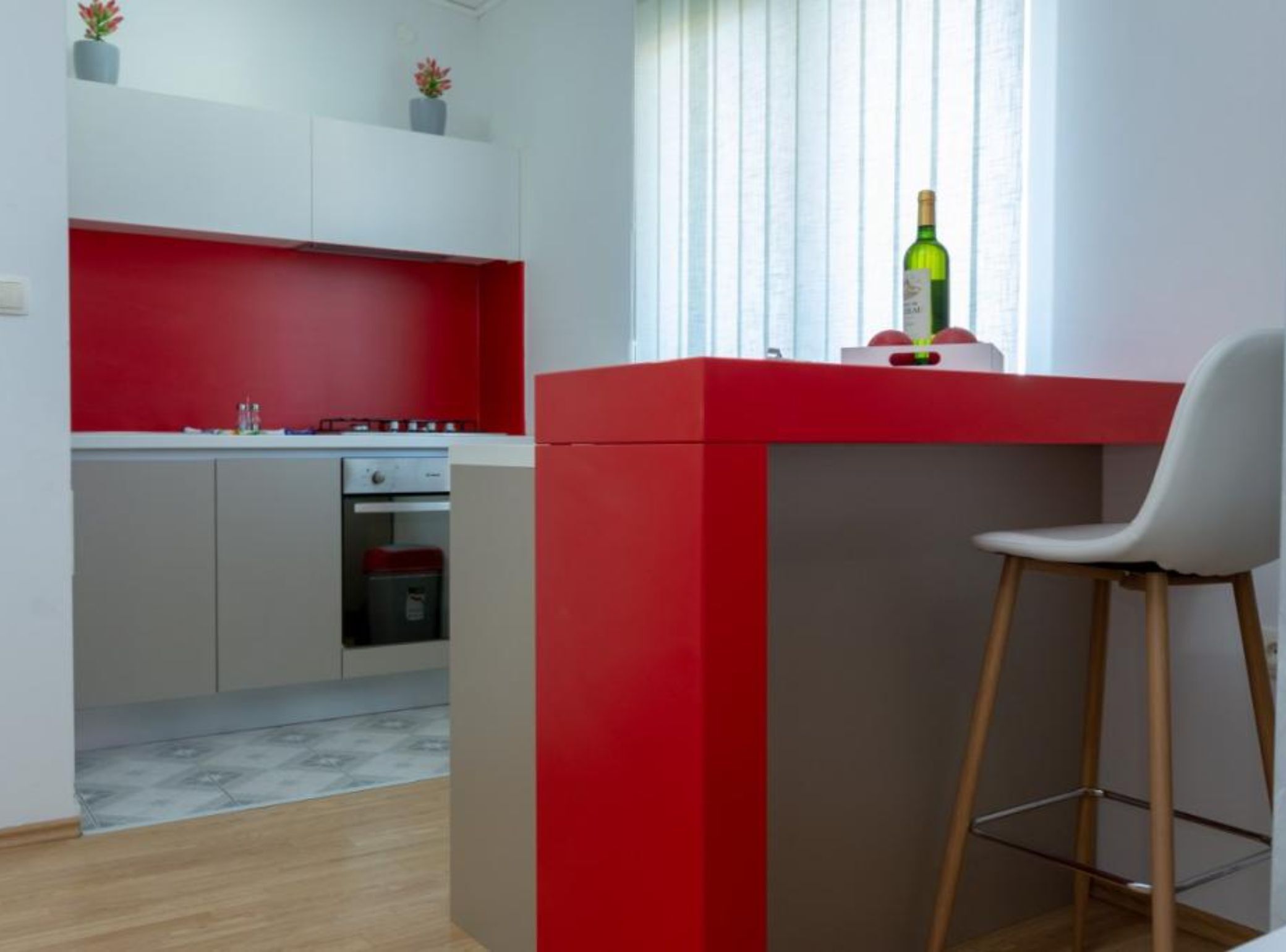 A compact kitchen with red and white cabinetry, a small dining area with a white chair, and a bottle of wine on the counter.