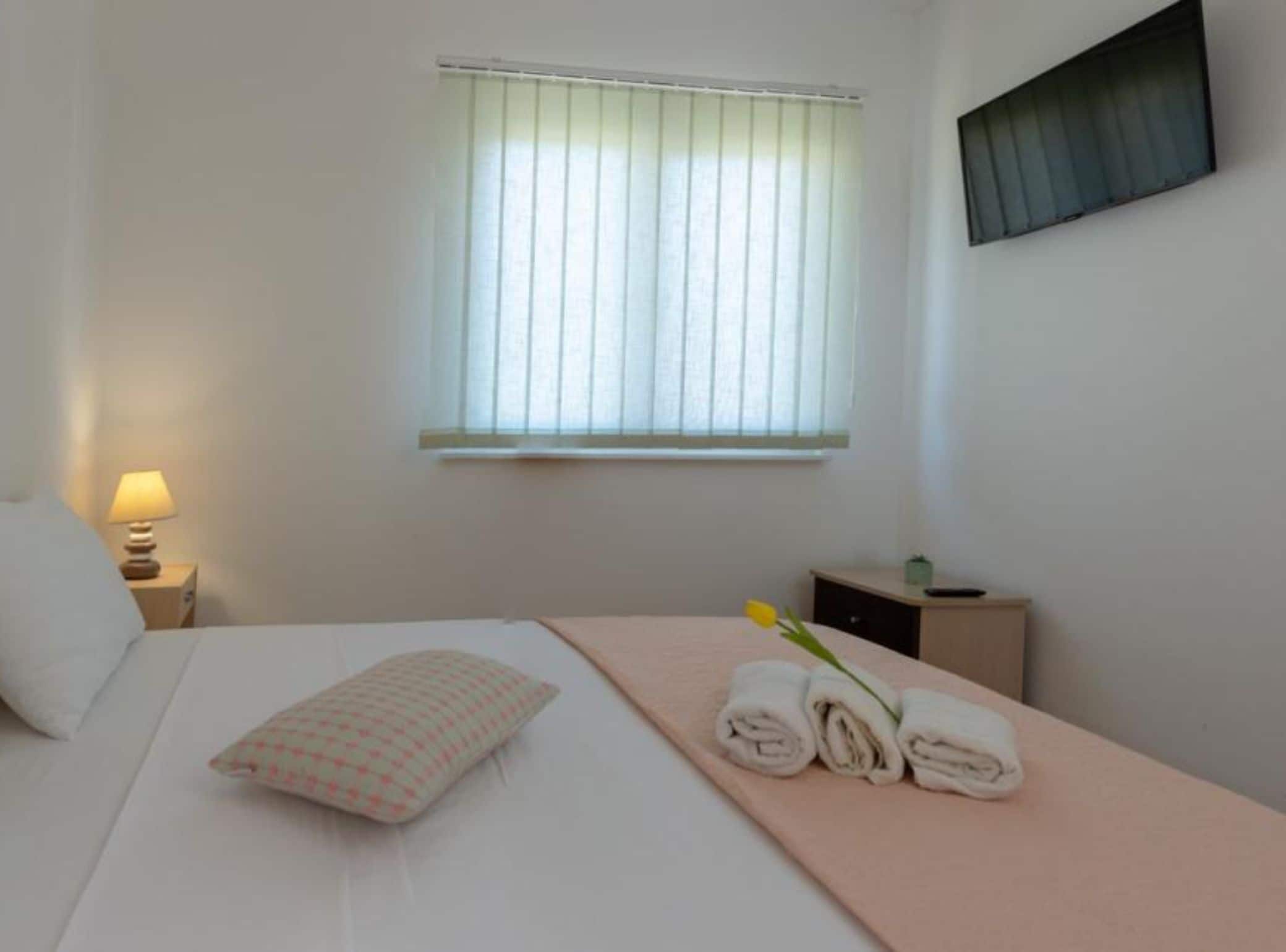 Hotelkamer met een bed, nachtkastje met lamp, raam met jaloezieën en een tafeltje met handdoeken en een bloem.