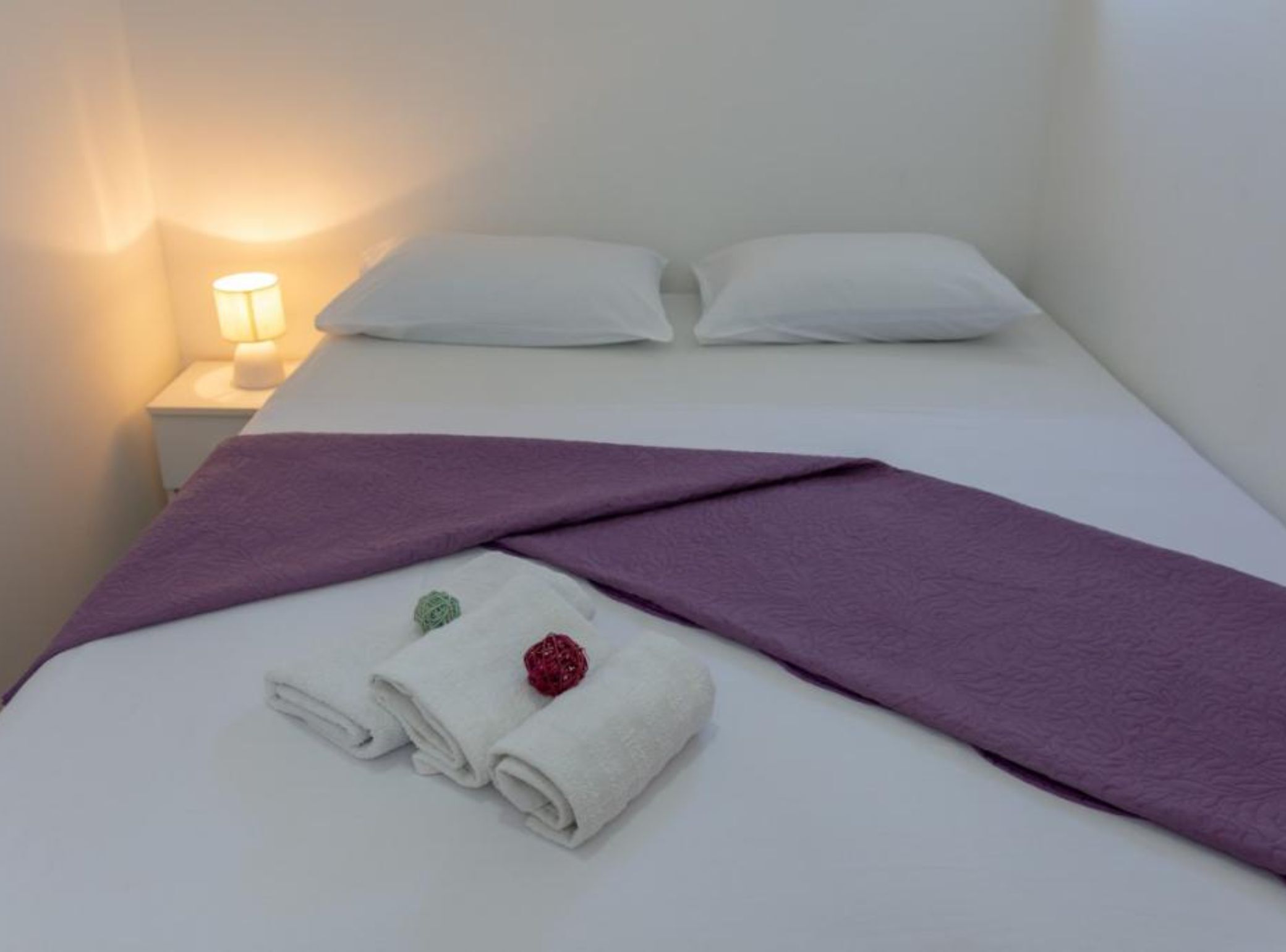 Ein ordentlich bezogenes Bett mit weißen Leinen, einer violetten Decke und zwei aufgerollten Handtüchern mit dekorativen Blumen.
