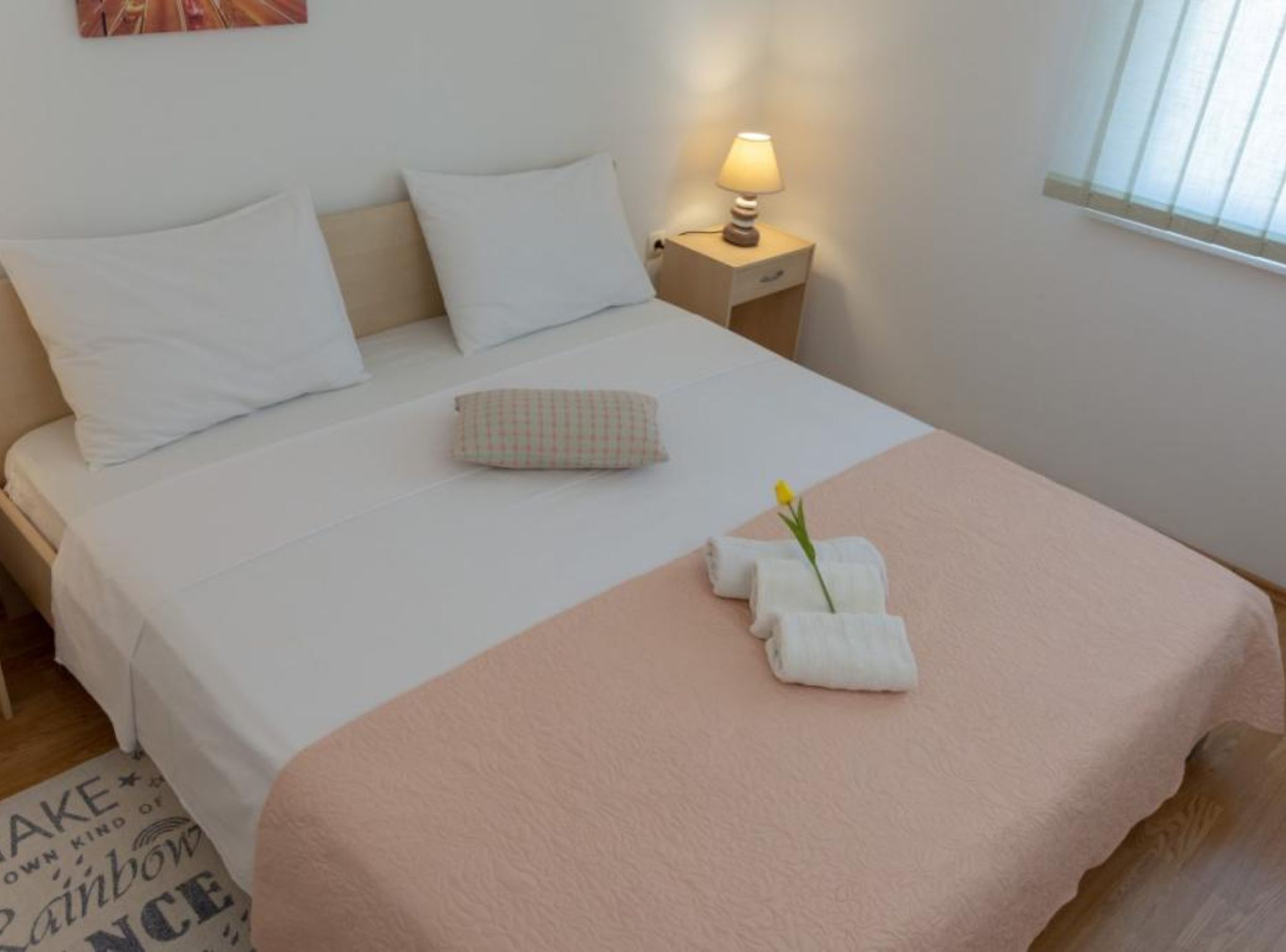 A neatly made bed with white and pink bedding, a small bedside table with a lamp, and a single yellow flower.