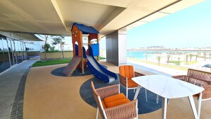 A covered outdoor area with a children's play structure, seating, and a view of the beach. - Beachfront Apartment (Lusail)