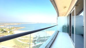 A balcony with a glass railing overlooking a coastal area with a clear view of the sea and distant land. - Beachfront Apartment (Lusail)
